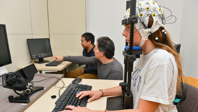 Students work in a psychology lab. One of them wears equipment on their head hooked to a monitor. 