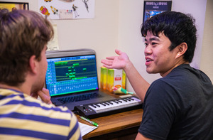 Two students sit at a desk in their residence hall room. They are working on a laptop that is displaying a music creation program.