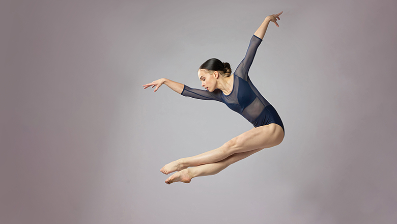 Female dancer in a pike jump, dark hair in tight bun, dark blue leotard with sheer 3/4-length sleeves.