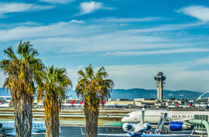 a long shot of LAX during the daytime. There are planes on the runways and palm trees in the foreground.