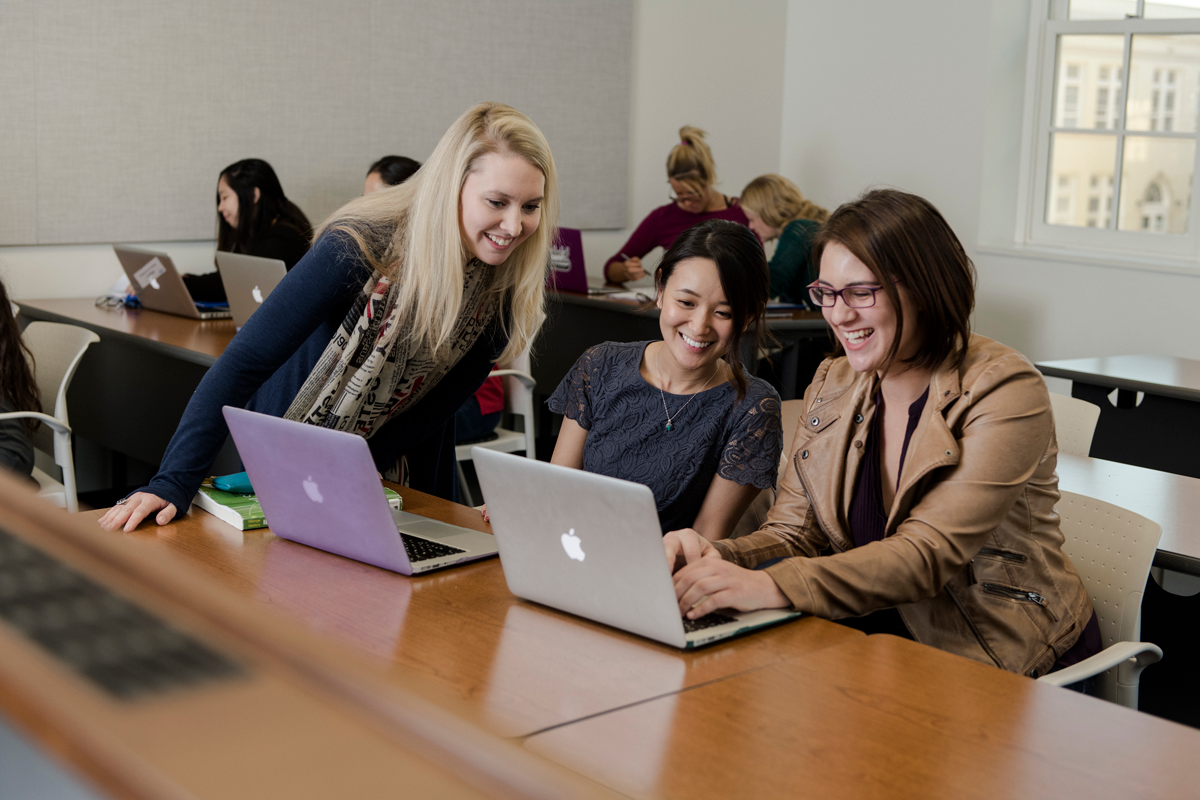 Two women sit at desk smiling at laptops in front of them while another woman leans over desk to see their screens