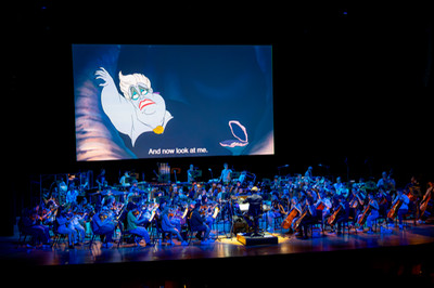 Orchestra performing on stage with The Little Mermaid movie playing on screen at rear of stage.