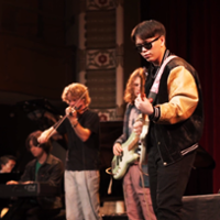 Student playing electric guitar on stage wearing sunglasses and varsity jacket.