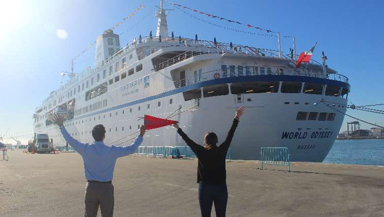 Two people holding a red flag facing a ship